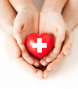 Child has their hands placed inside of the palms of an adult forming a heart in hands. The child is holding a heart shaped object with a cross on it