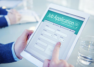 A male is sitting at a table with a cup of water next to him and a tablet in his hands that has a Job Application tab open